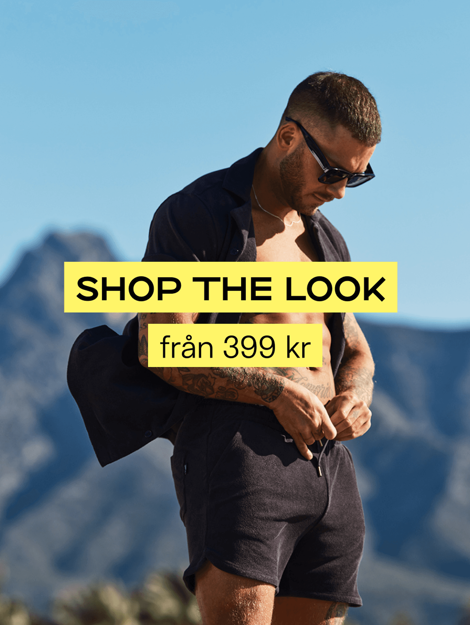 Man in sunglasses wearing a dark outfit adjusts his shorts outdoors against a mountain backdrop. Text: "SHOP THE LOOK från 399 kr."